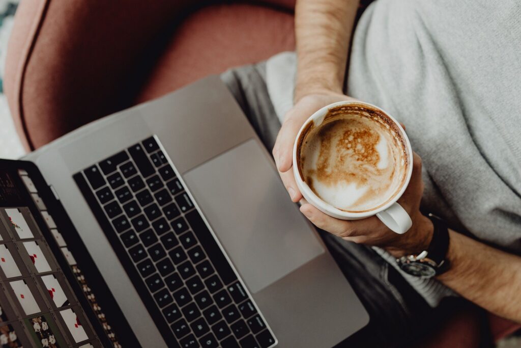 Ein Mann sitzt mit einer Kaffeetasse vor einem Laptop. Entspannt kann man von zu Hause oder aus dem Büro Onlinecoaching nutzen.